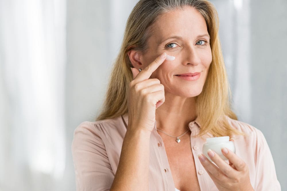 Smiling senior woman applying anti-aging lotion to remove dark circles under eyes