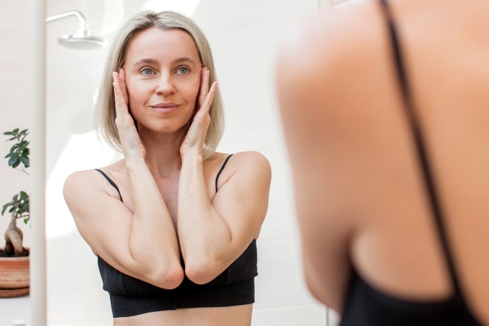 Middle aged woman is thinking about facelift. 40s woman looks in the bathroom mirror at home