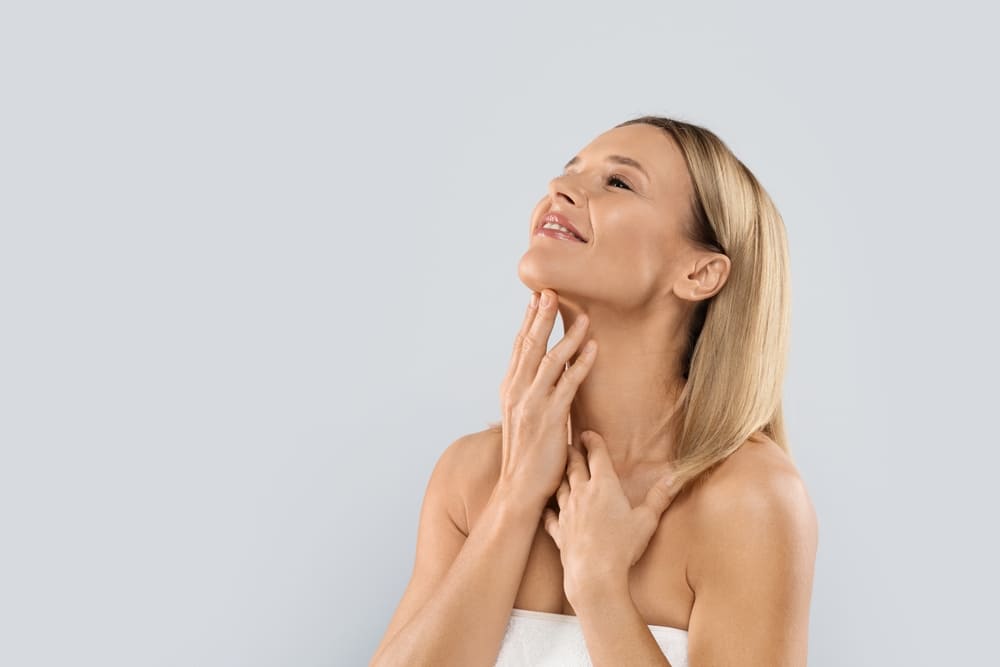 Smiling beautiful half-naked middle aged blonde woman covered in white towel touching her neck,