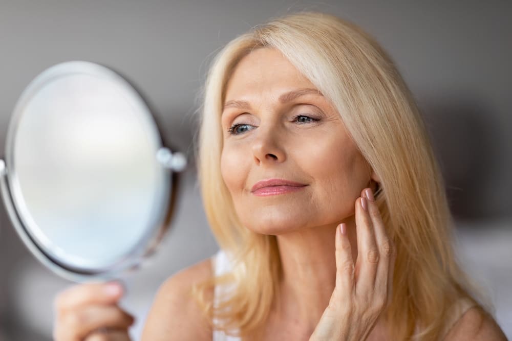 Attractive aged european woman looking in magnifying mirror and touching her face smooth skin