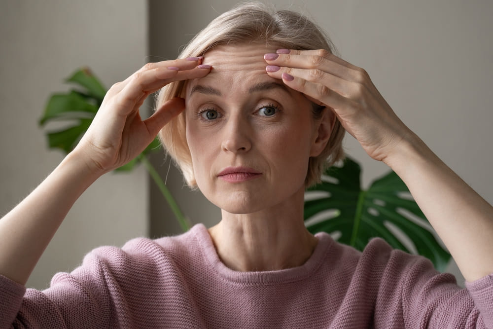 mature woman is captured indoors while gently touching her forehead with both hands, appearing introspective or concerned