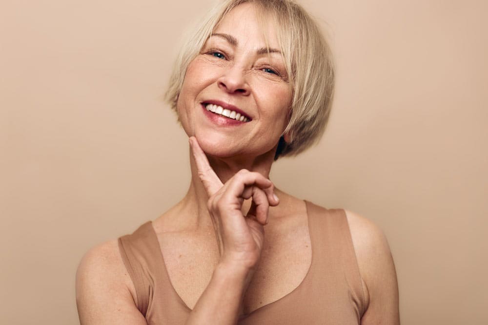 Smiling middle aged woman with short blonde hair touches chin gently.